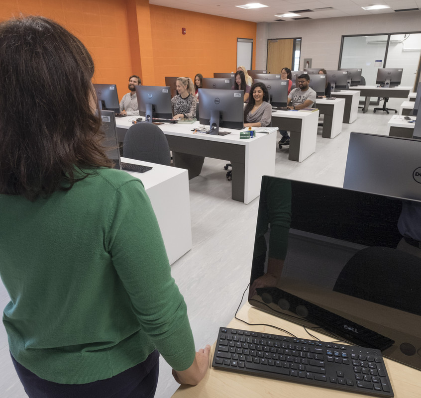 A teacher looks at her class of students, sitting at their computer stations.
