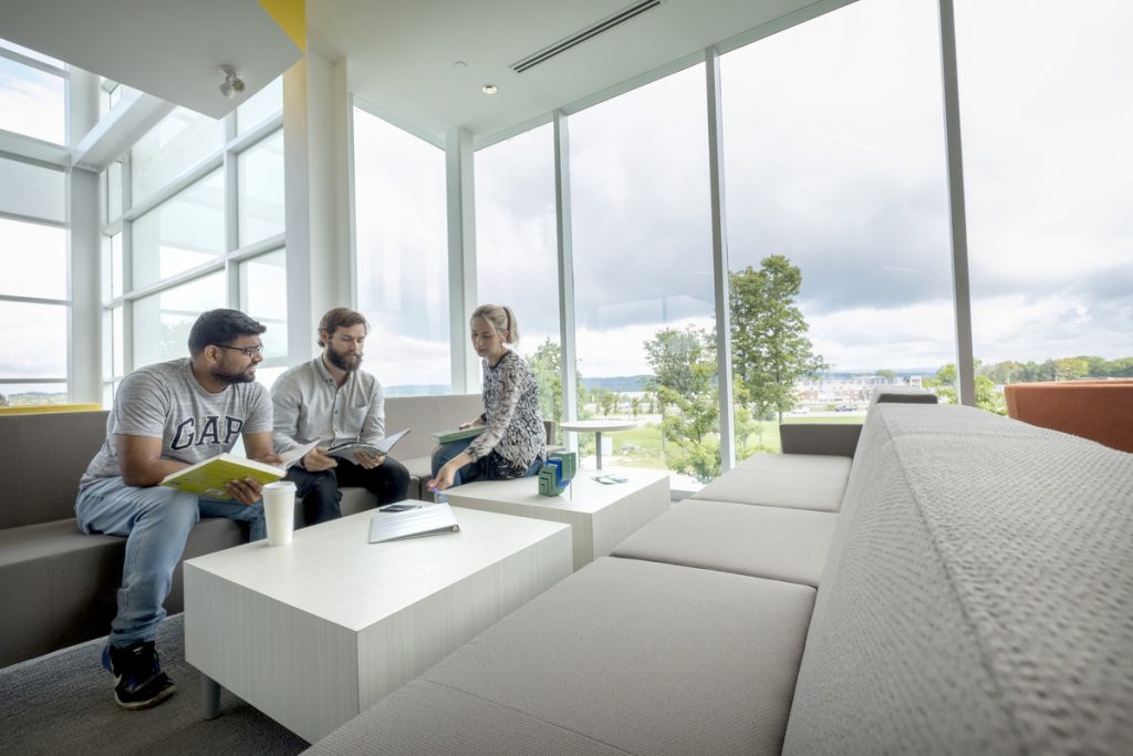 Three students sit on a couch and table in a student lounge with floor to ceiling windows.
