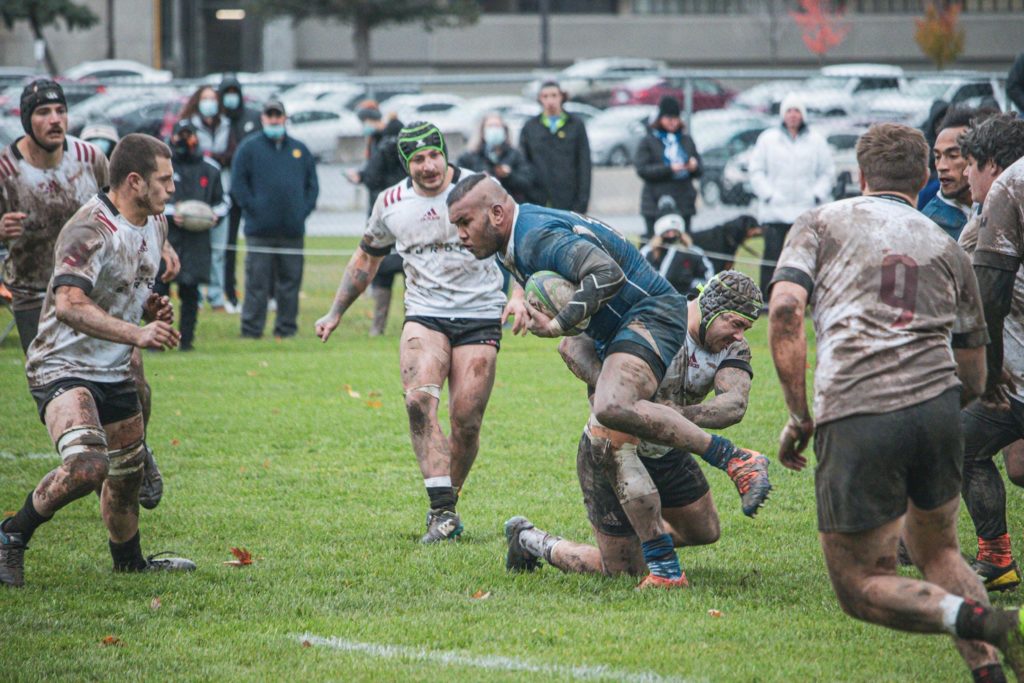 A group of muddy rugby players during a game, with one in the centre gripping the ball and running past players on the opposing team.