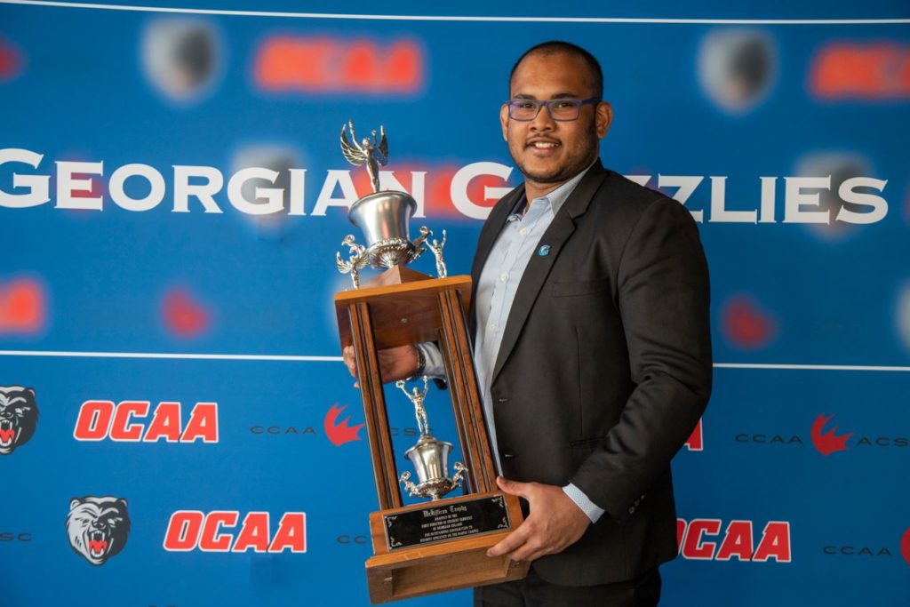 A person wearing glasses and a suit smiles and holds up a rugby trophy. The backdrop behind them reads Georgian Grizzlies.