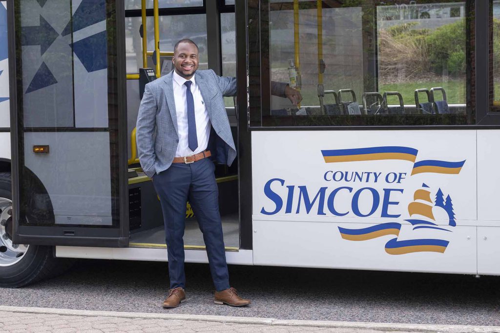 Ronjay Clarke in a suit and tie stands in front of a County of Simcoe bus