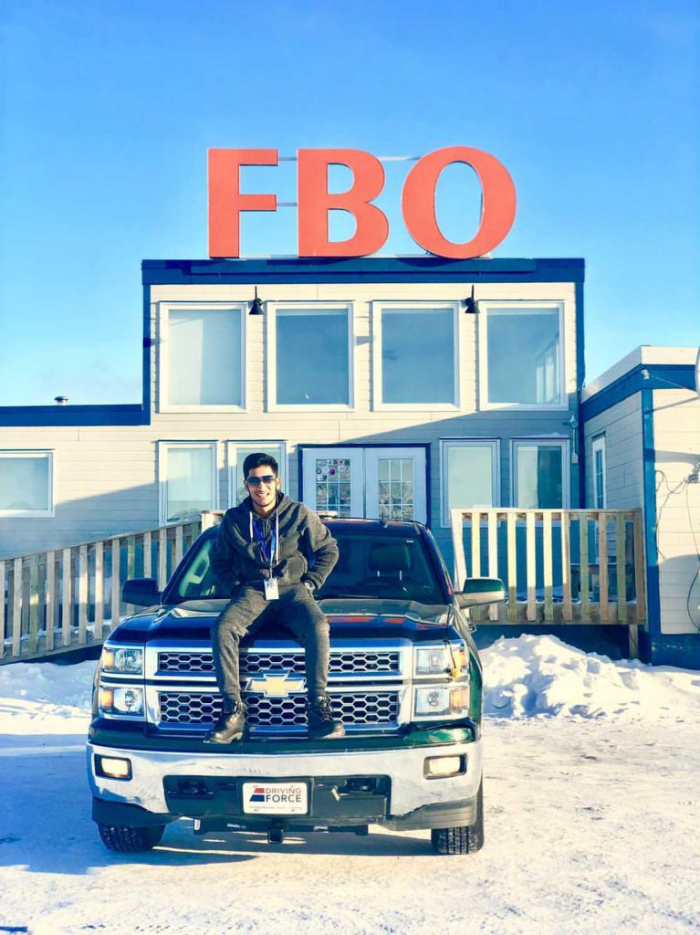 A person with short, brown hair and wearing a dark sweater, pants, boots and sunglasses, sits on the hood of a black pickup truck parked on snow in front of a building with a sign reading FBO.