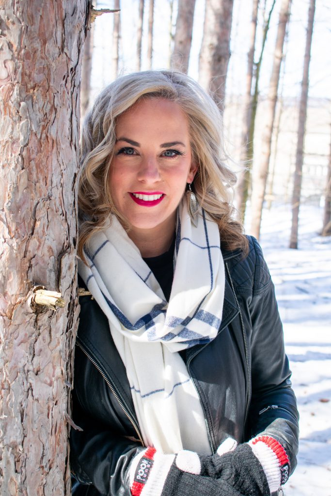 A person stands outside and leans against a tree in a snowy forest and smiles at the camera.
