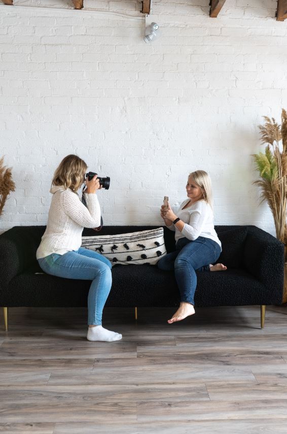 Two people sit on a couch and face each other, while one takes a photo of the other.