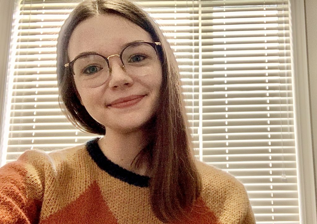 A selfie of a person with long, brown hair, glasses and an orange and yellow sweater.