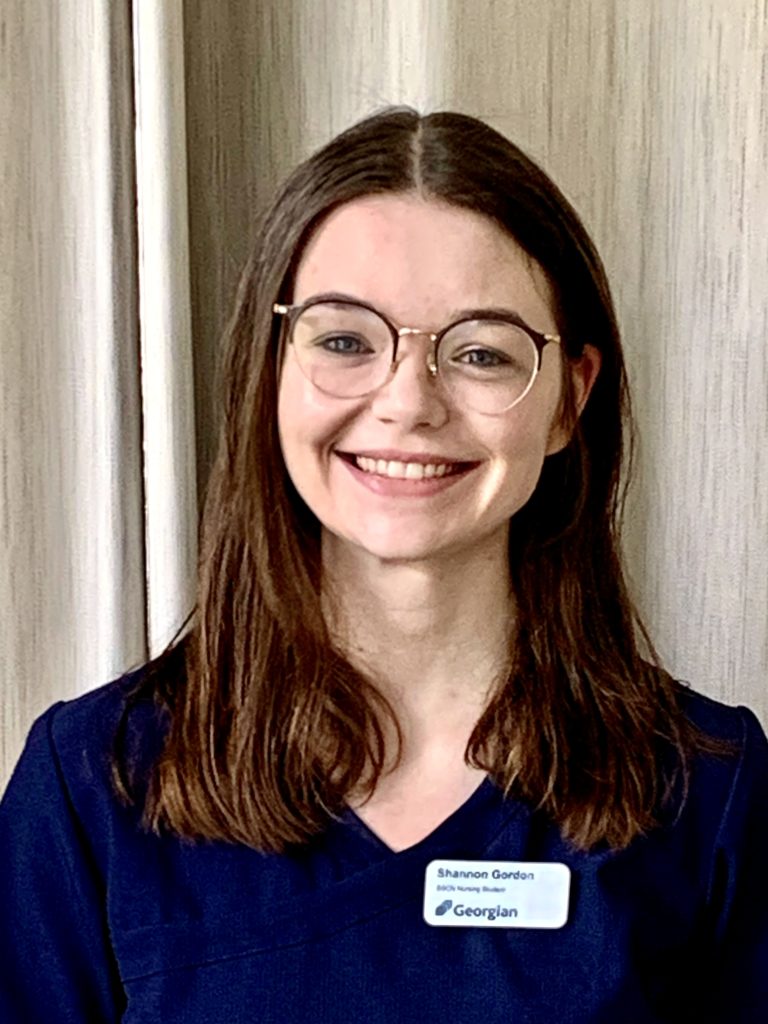 A headshot of a person with long, brown hair, glasses and blue nursing scrubs with a nametag reading "Shannon Gordon, BScN nursing student, Georgian College."