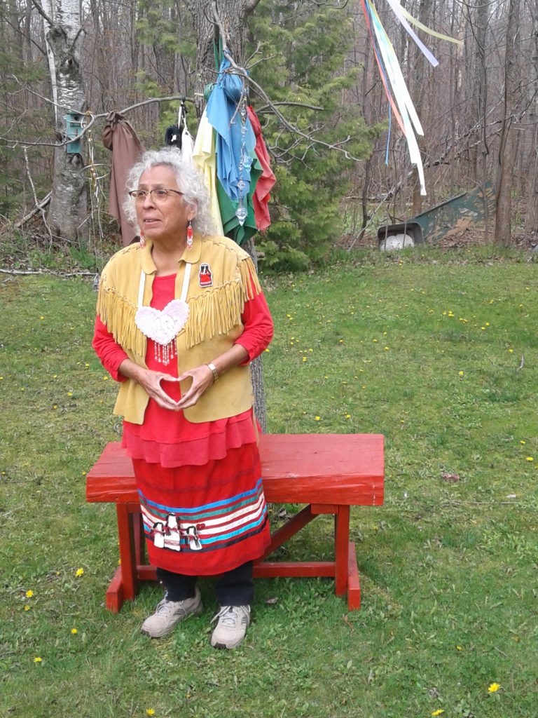 A person wearing black pants, red skirt and shirt, and fringed brown vest stands outside on grass near a wooded area.