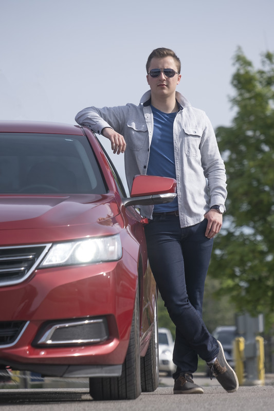 A person with short brown hair, dressed in jeans, blue shirt and grey jacket, and sunglasses, looks at the camera and leans an arm against a red car.