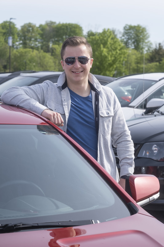 A person with short brown hair, sunglasses, blue shirt and grey jacket, smiles and leans an arm against the side of a red car.
