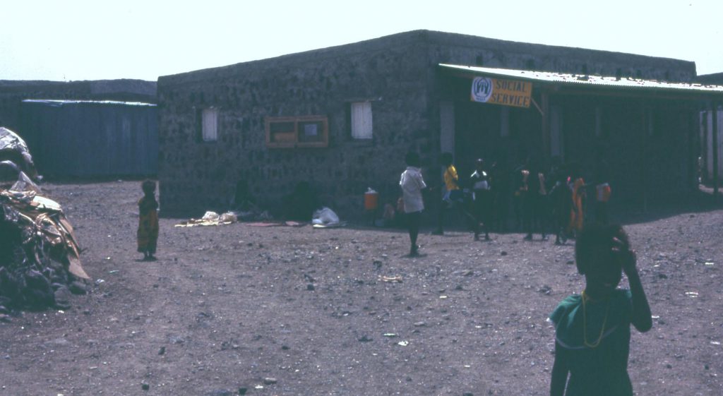 People stand around outside of a building in an African village.