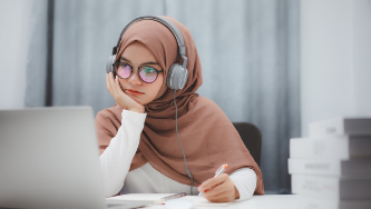 student wearing a brown hijab and headphones looking at a laptop screen and taking notes with a pen