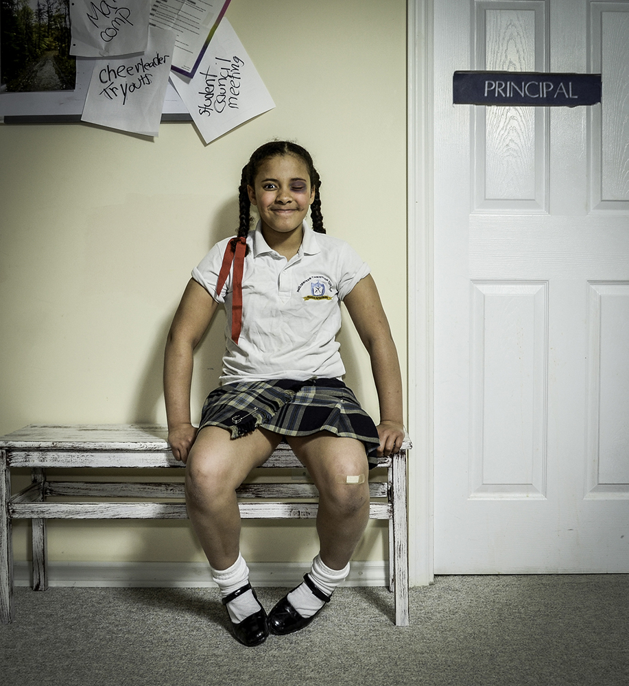 Elementary aged girl sitting with pigtails with a bruised eye outside of the principal's office