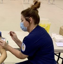 A side view of a person in blue scrubs and face mask giving someone a vaccination injection in their arm.