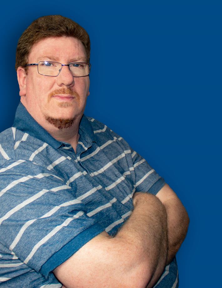 A person with short auburn hair, glasses and a blue and white striped shirt, against a blue background, crosses their arms and looks at the camera.