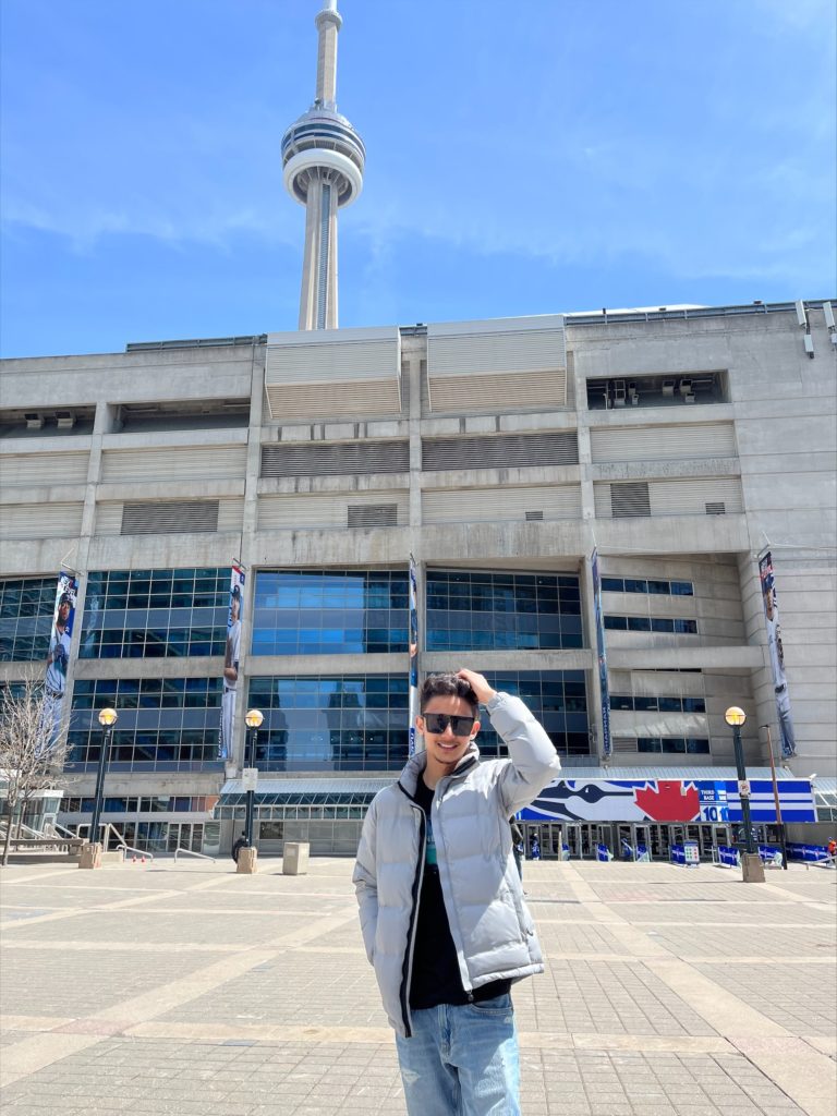 A person with brown hair, black sunglasses, grey jacket and jeans stands in front of a large building and tower.