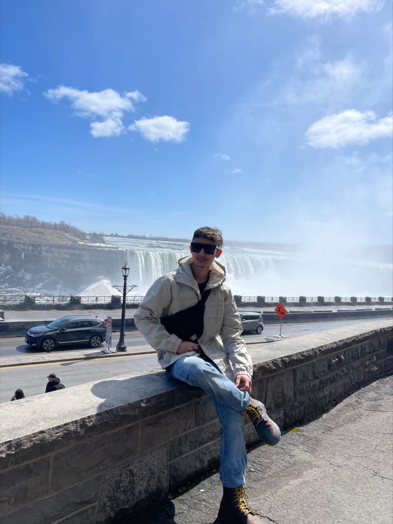 A person with short brown hair, black sunglasses, tan jacket, black shirt, jeans, and brown boots, sits on a short brick wall in front of Niagara Falls, a large waterfall.