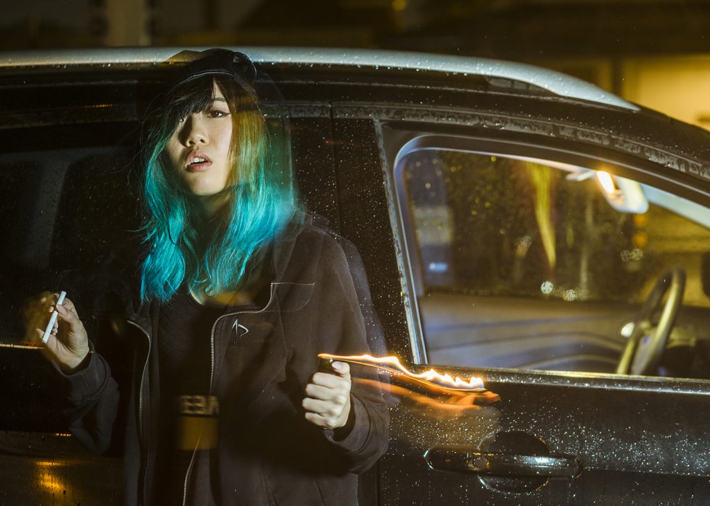Asian woman with blue hair standing outside an SUV in the rain smoking a cigarette