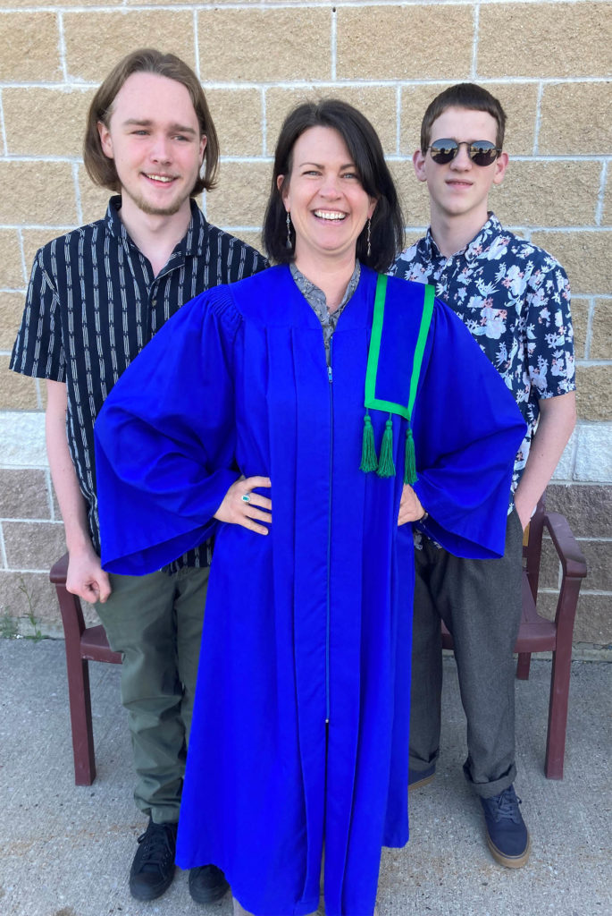 A female with dark hair wearing a blue graduation gown standing outside against a brick wall. She is flanked by two young males.