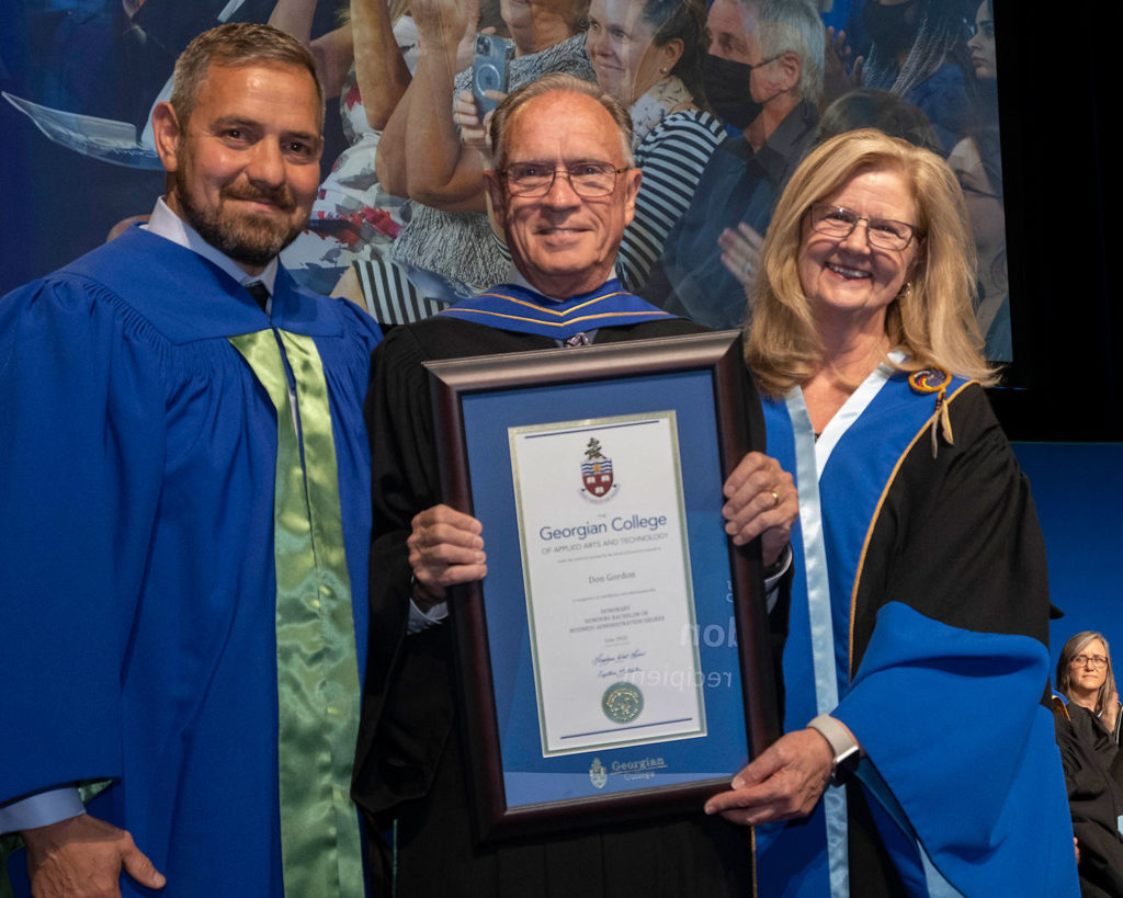 An older white man with glasses holding a framed certificate. He is flanked by a blonde white woman on the right and a white man on the left. They're all wearing formal graduation gowns.