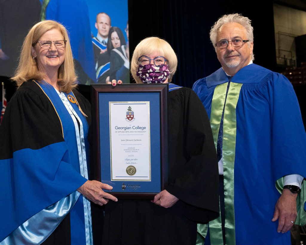 A blonde white woman wearing a face mask and holding a framed certificate. she is flanked by another blonde white woman on the left and an older white man on the right. They are all wearing formal graduation gowns.