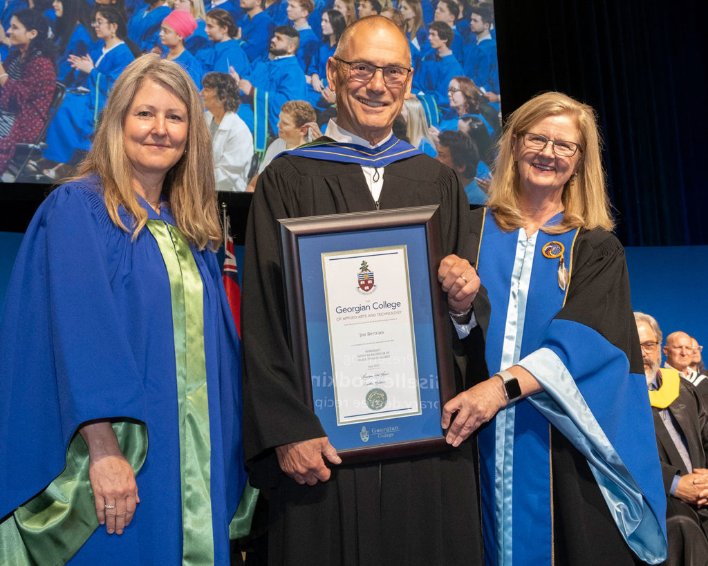 An older white man with glasses holding a framed certificate. He is flanked by a blonde white woman on the right and another white blonde woman on the left. They're all wearing formal graduation gowns.