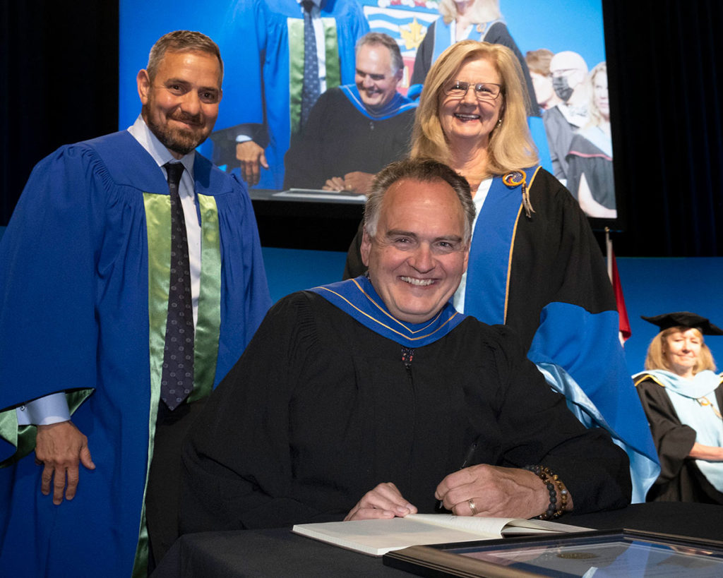 A white man with grey hair sitting at a table and smiling. There is a smiling white man and a blonde white woman standing behind him.. They're all wearing formal graduation gowns.