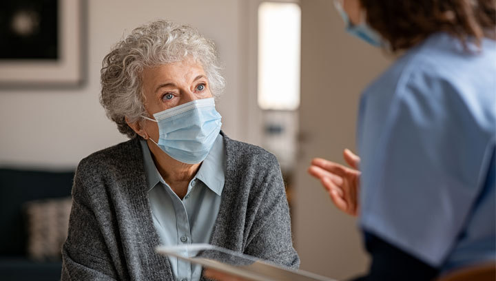 An elderly person with gray, curly hair wearing a mask next to a PSW wearing scrubs, a mask and holding a tablet