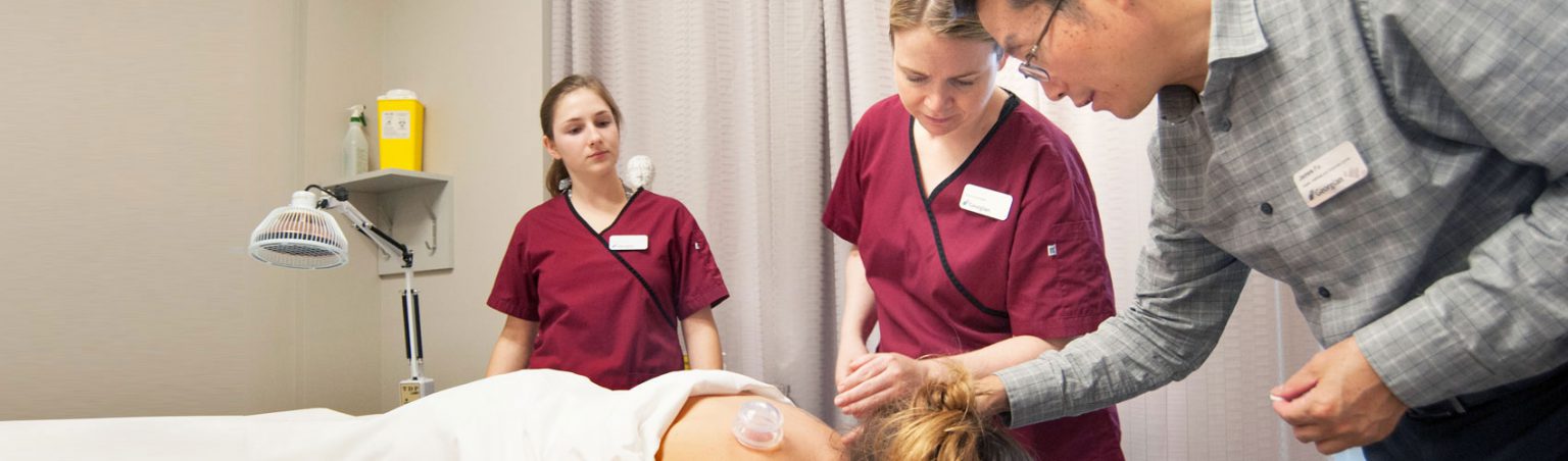 Two acupuncture students in scrubs watch an instructor apply cupping therapy to the upper back of a patient who is laying down