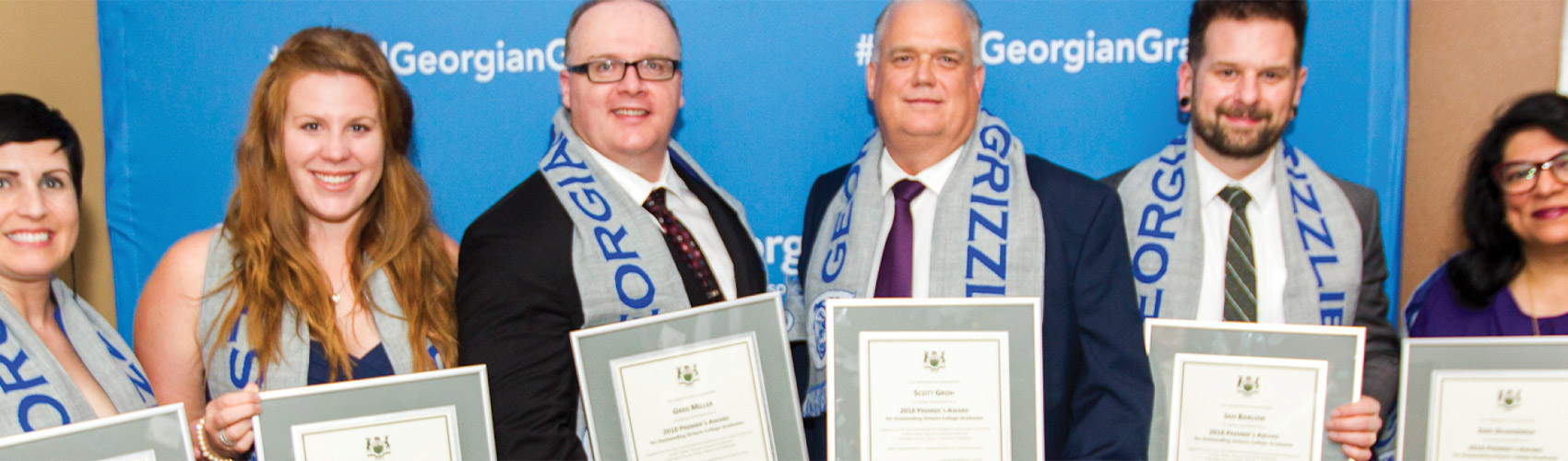 A group of people holding framed certificates while wearing gray and blue "GEORGIAN GRIZZLIES" knit scarves around their necks