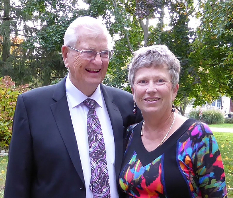 An older white couple standing outside smiling at the camera. They're dressed up in business attire.