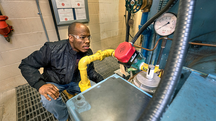 Person wearing plastic safety glasses crouching down next to a piece of machinery in the skilled trades lab at Georgian's Owen Sound Campus