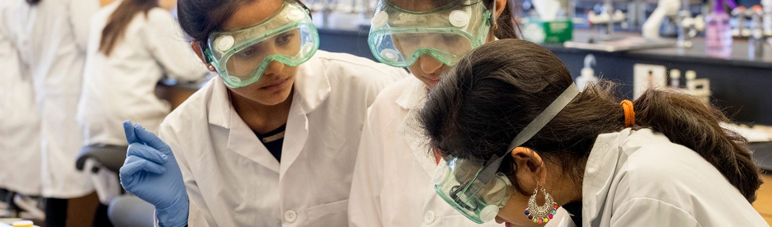 Three students wearing white lab coats, safety goggles and blue latex gloves working with biological samples in a science lab