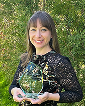 A smiling white female with long dark hair standing outside holding up a glass award
