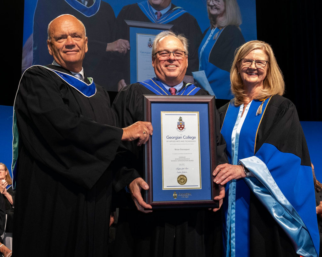 An older white man with grey hair and glasses holding up a framed certificate. He's flanked by a blonde white woman on the right and a white man on the left. Everyone is wearing formal graduation gowns and smiling.