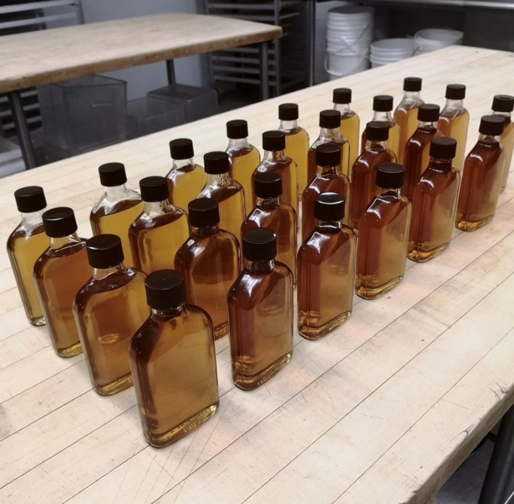 About 30 bottles of maple syrup sit in glass bottles on a kitchen counter.
