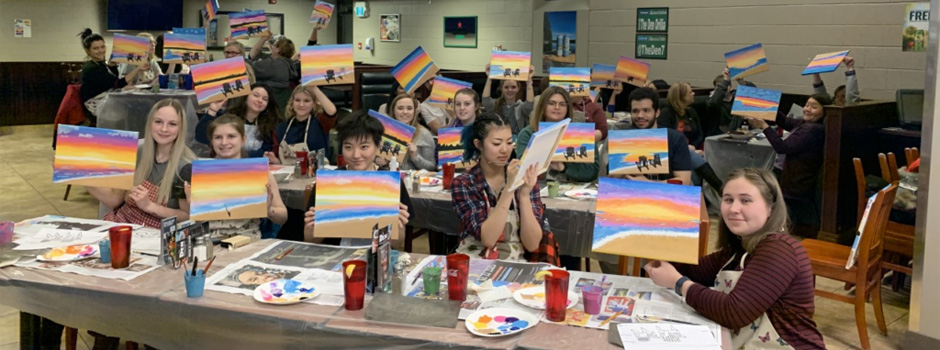 Students sitting at tables with art supplies holding up canvasses painted with a scenic beach
