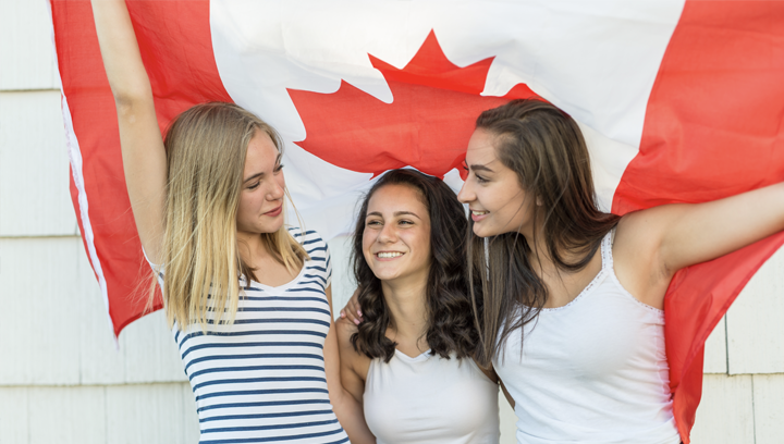 Three women standing close together smiling under a Canadian flag. 