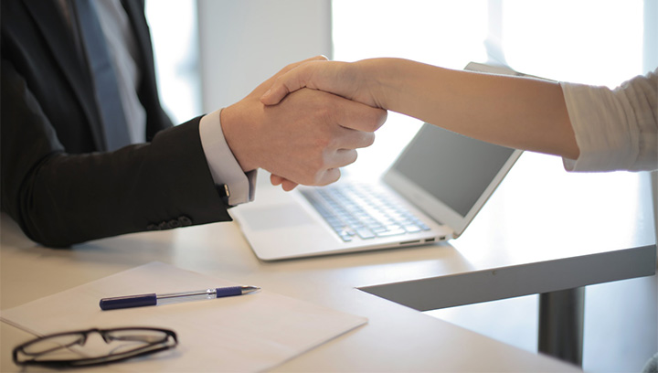 Two people in business attire shaking hands, featuring a laptop on a table in the background