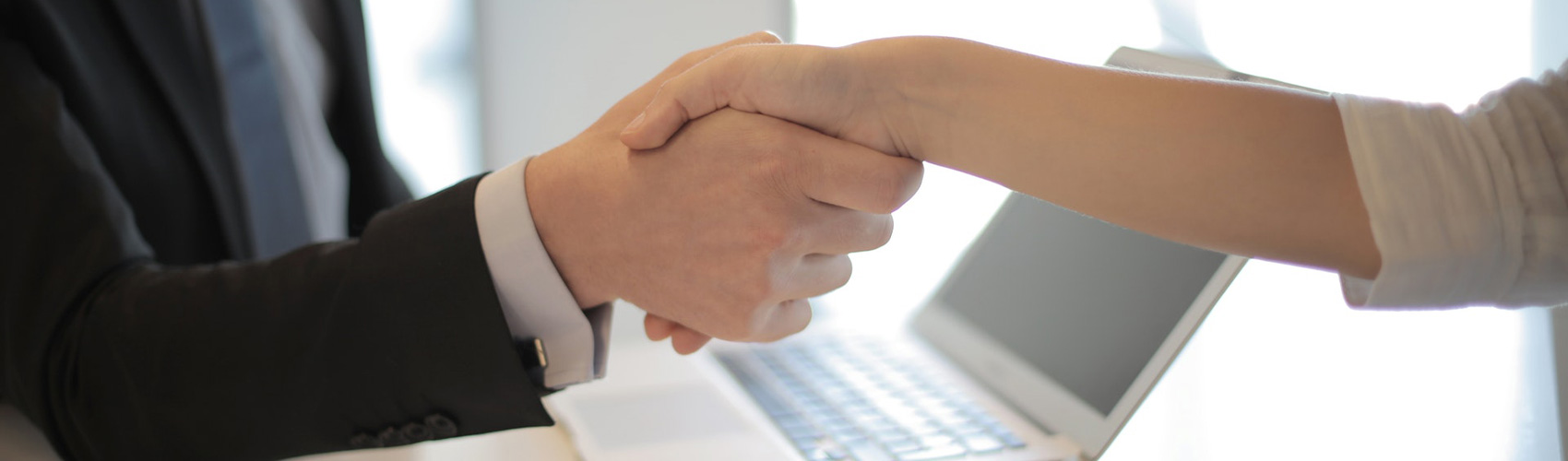 Two people in business attire shaking hands, featuring a laptop on a table in the background
