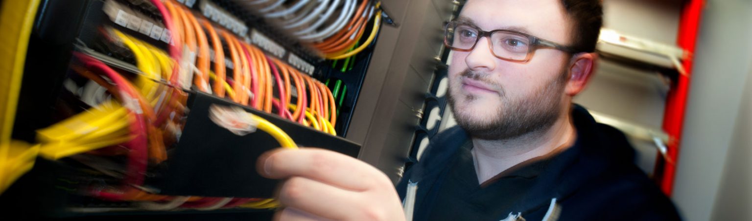 A network technician with brown hair, speckled glasses and a navy blue zip-up sweater connecting a wire to server equipment