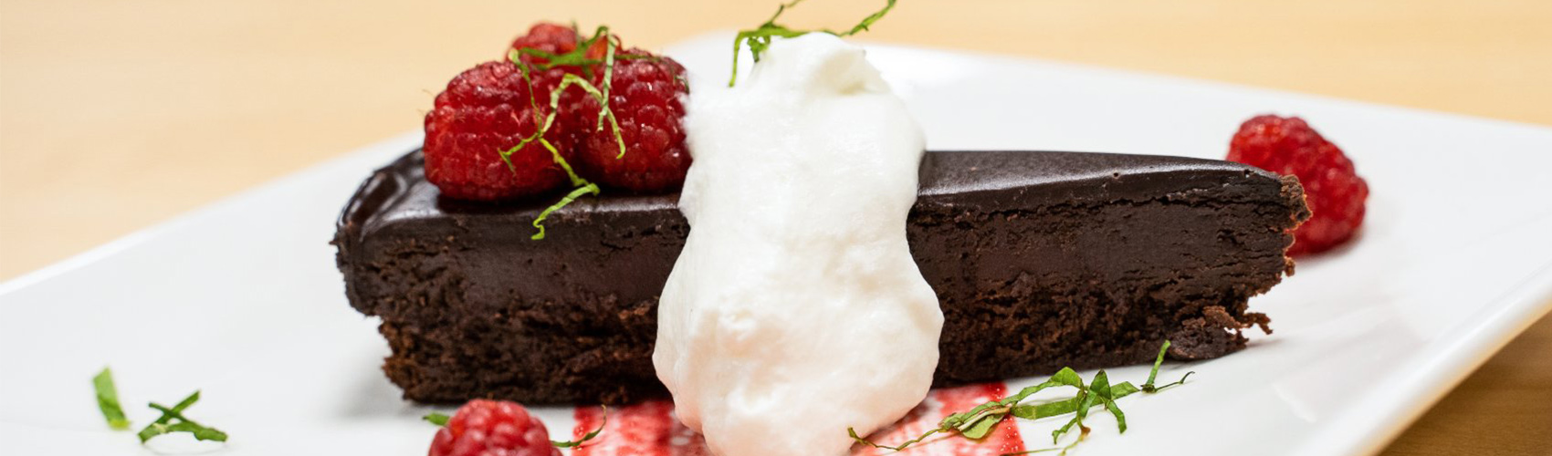A slice of chocolate cake garnished with whipped cream and raspberries on a white square plate