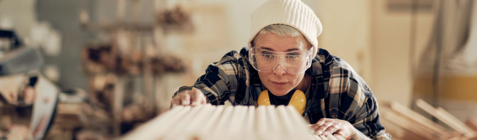 A person in a plaid shirt, white toque, wearing clear safety goggles with earmuffs around their next, looking at a piece of lumber in a shop setting