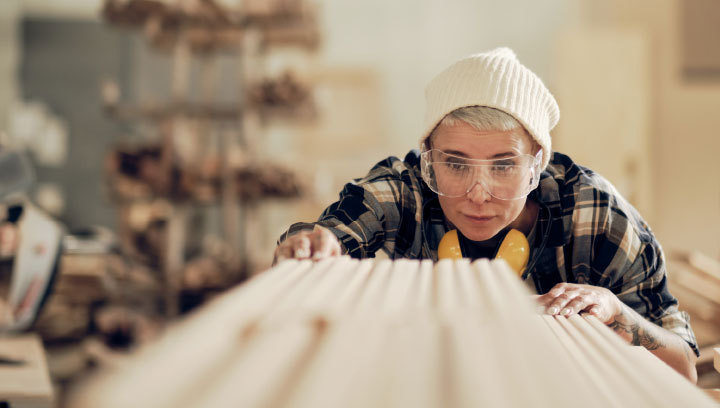 A person in a plaid shirt, white toque, wearing clear safety goggles with earmuffs around their next, looking at a piece of lumber in a shop setting