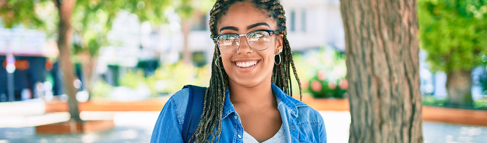 Person wearing glasses, hoop earrings and a denim shirt smiling and looking off to the side in an outdoor setting