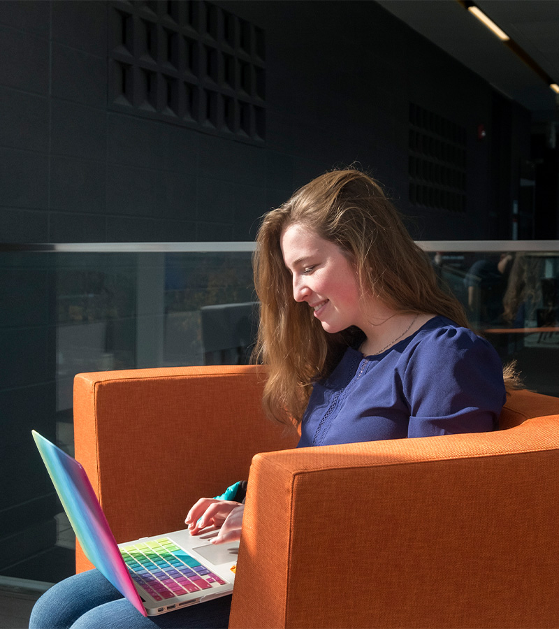 female student sitting on an orange lounge chair using a Mac book laptop