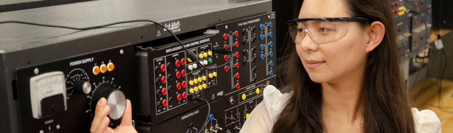 Electrical engineering professional with dark hair and safety goggles turning a power supply dial on a piece of equipment