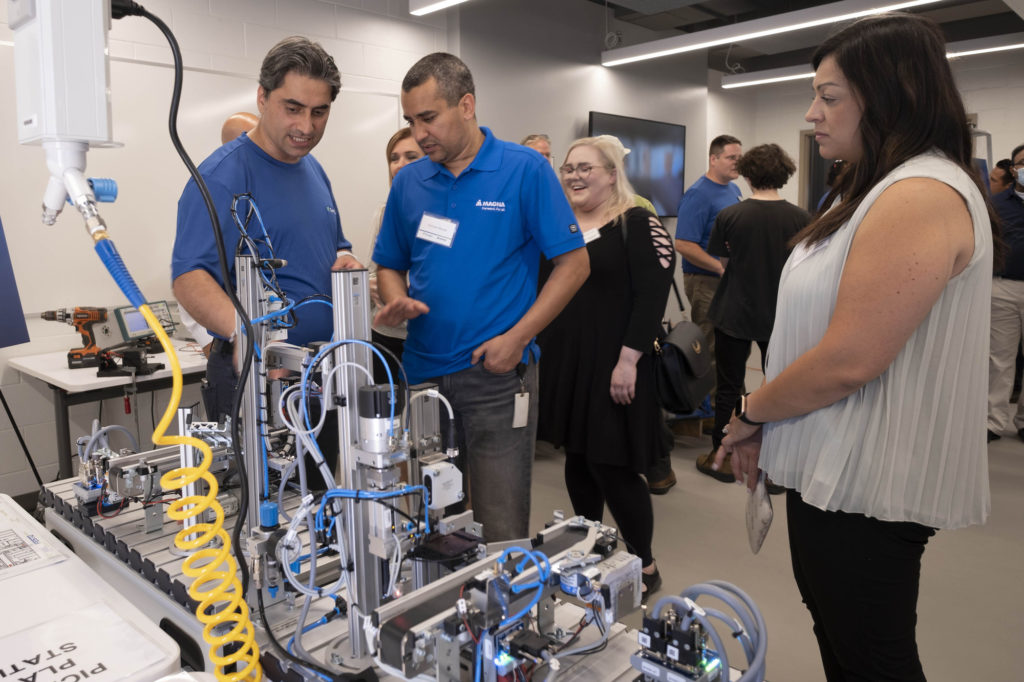 Two males in blue t-shirts explaining mechatronics equipment to a female.