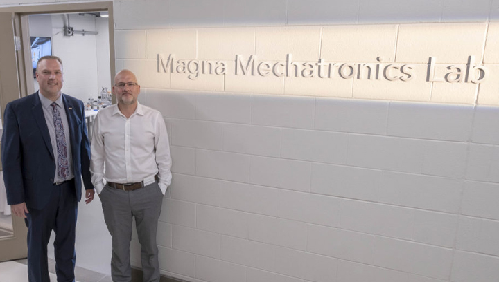 Two white males in business attire standing in a hallway beside a wall that has a sign saying Magna Mechatronics Lab