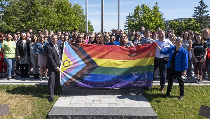 A group of Georgian employees holding a large pride flag in front of the Barrie Campus Cenotaph prior to the flag raising for Pride Month 2022