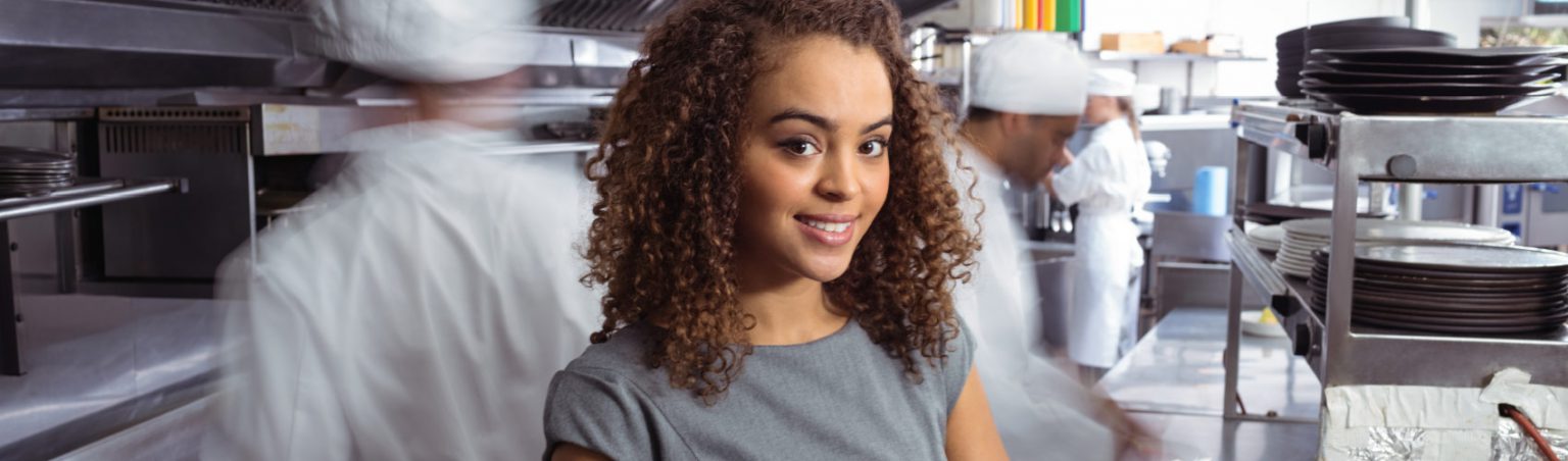 An event manager with brown, curly hair in a heather gray dress taking notes on a clipboard inside a commercial kitchen